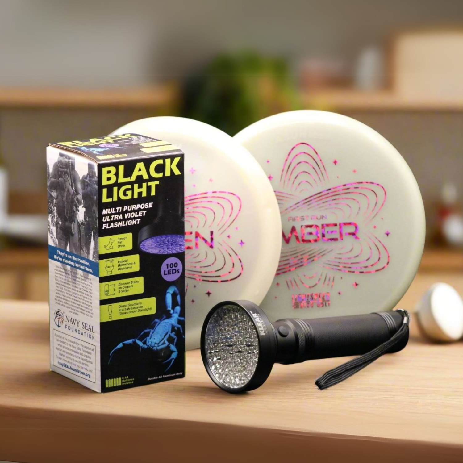 Black light UV flashlight with packaging displayed in front of two Lightspeed Disc Golf glow-in-the-dark Ember discs on a wooden table.